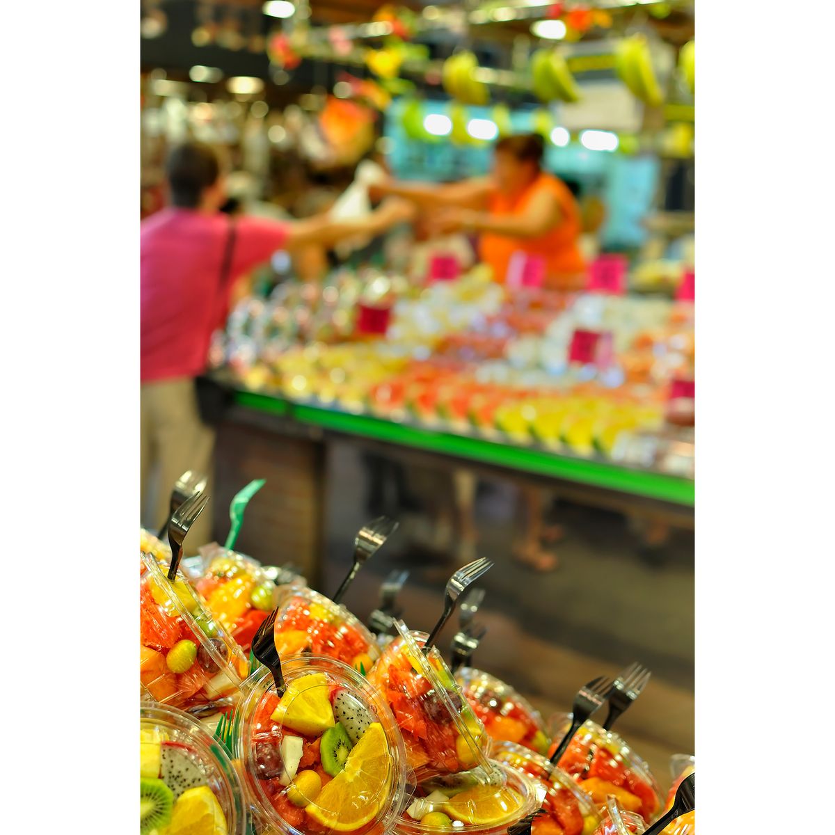 Fruit salads in the foreground with blurred background showing person paying for items in Barcelona&rsquo;s historic La Boqueria market