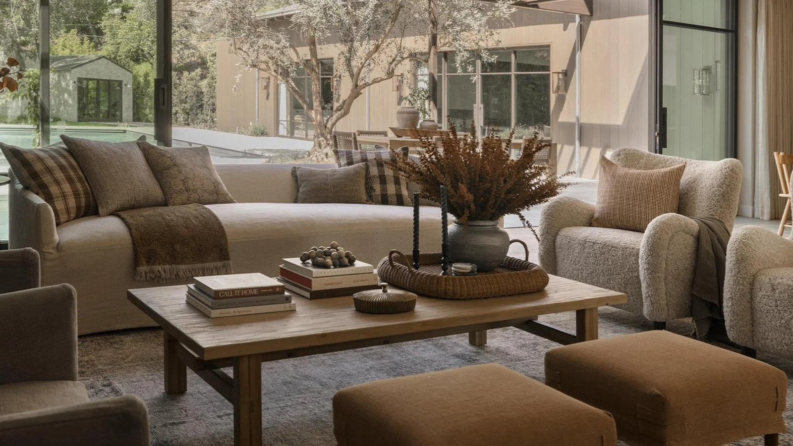 a cozy, neutral toned living room with a couch with many pillows, a coffee table with decor on top, two ottomans, and a wall of sliding doors that opens up to the backyard