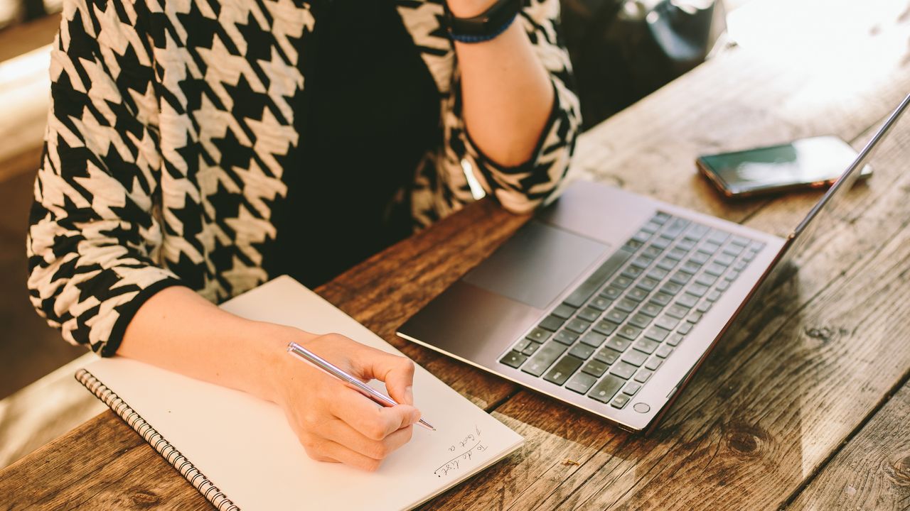 A woman writes on a pad of paper at her desk while sitting in front of an open laptop.