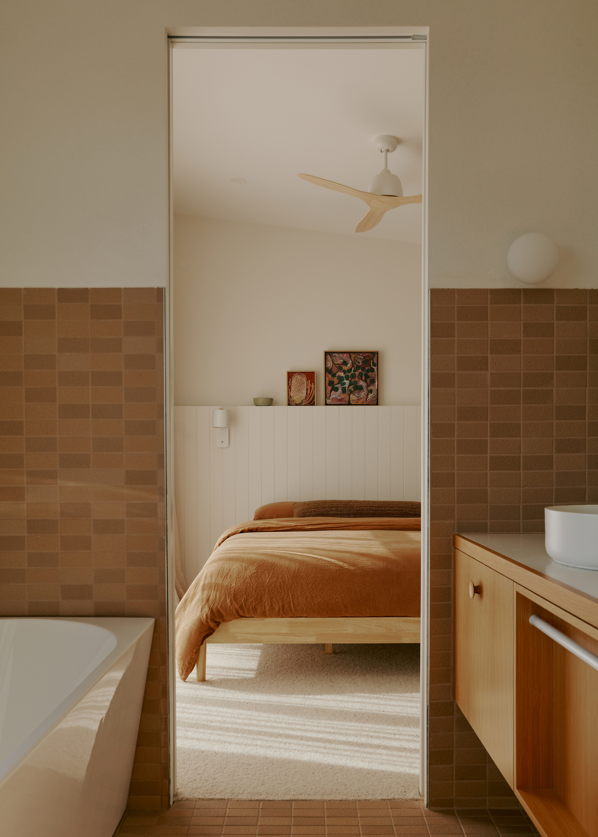 A bathroom leading into a white bedroom with a wooden bed frame, a tan bedscape, a ceiling fan, a panelled wall with sitting paintings