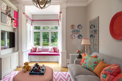 family room/playroom with window seat, footstool, sofa, bright pink trimmings, tv, toy storage, bright cushions, pink and white rug