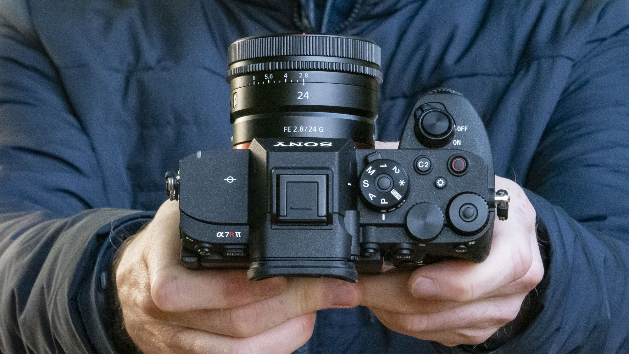 Sony A7R VI camera with 24mm F2.8 lens attached, in user's hands and they are pointing it up to show the top plate