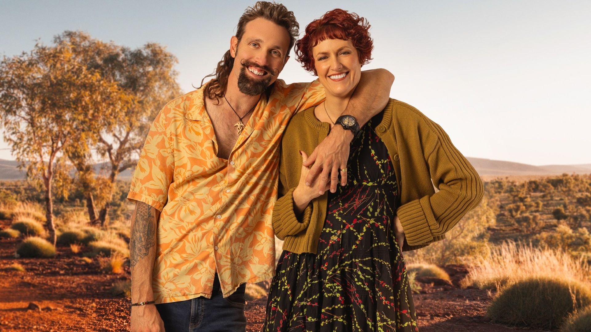 Andi and Nigel side by side in Australia. He has his hand over her and is in a button up vibrant shirt. She's wearing a dress with a sweater. They're both smilling. He has a mullet.