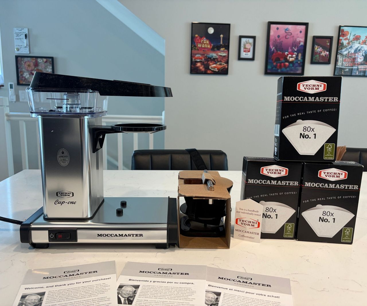 A silver Moccamaster cup one coffee maker, on a white counter beside a stack of three packs of filters.