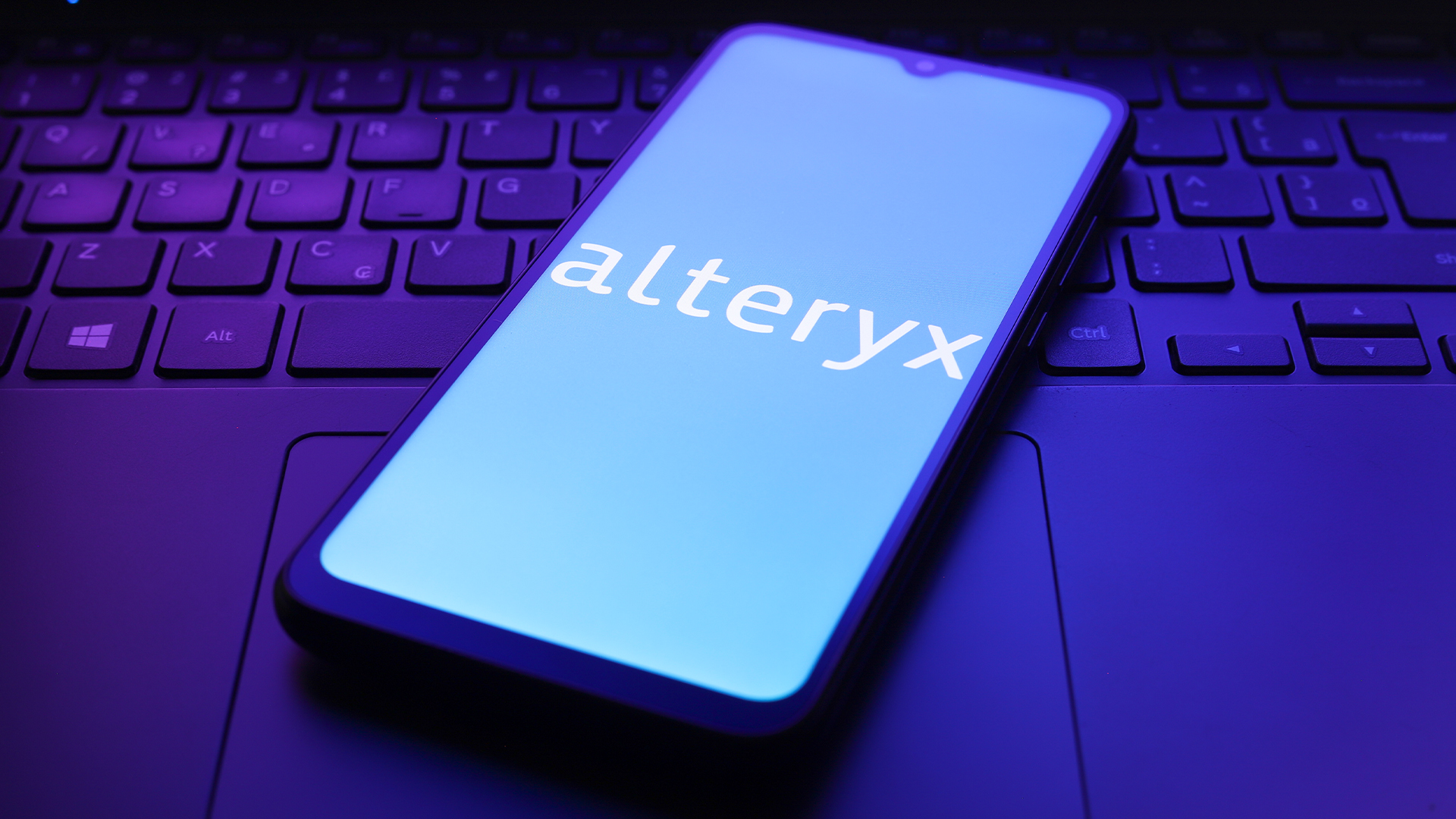 Alteryx logo and branding pictured on a smartphone screen placed on top of a laptop keyboard and mousepad. 