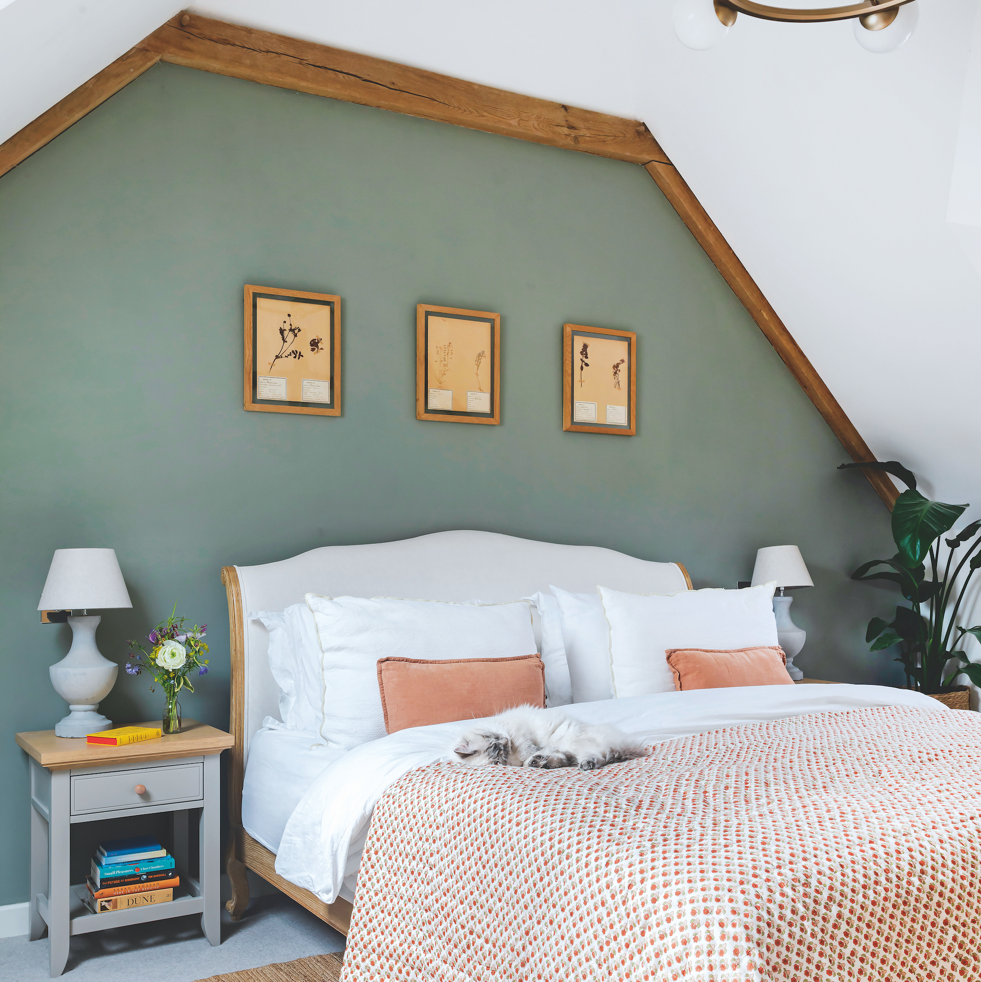 bedroom in oak frame house with a green painted wall with three pictures on it behind an upholstered bed with a printed quilt cover