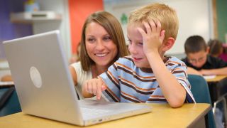 Young boy works on laptop computer while teacher looks over his shoulder smiling
