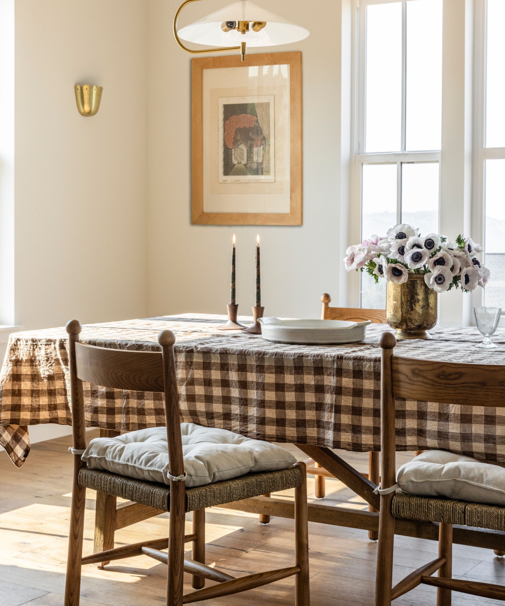 Dining room with a wooden table covered in a gingham tablecloth and vintage wooden chairs tucked underneath, metal candelsticks and vase with flowers and a framed print in the background