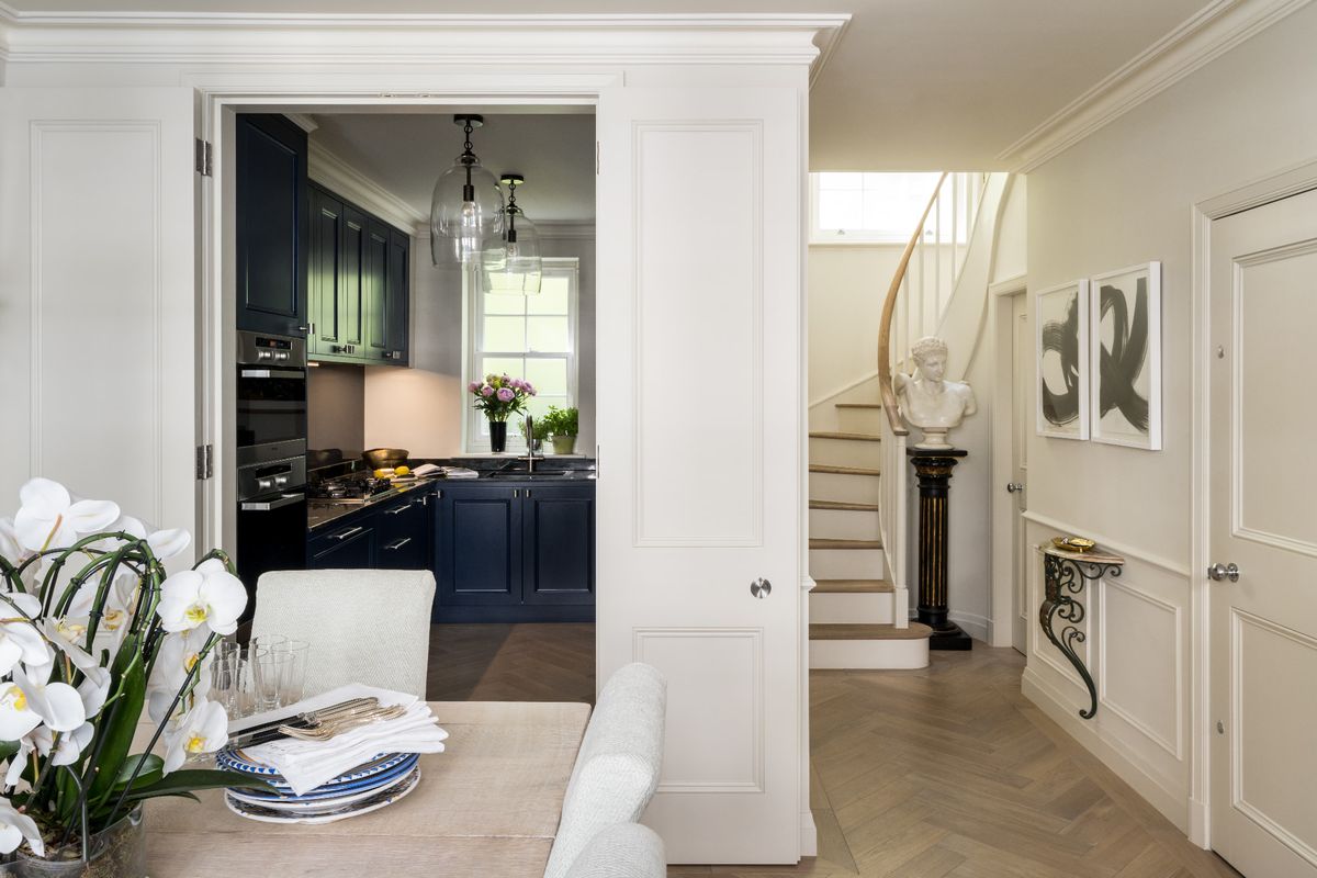 A white panelled room with light parquet flooring, a dinner table at the forefront and a blue kitchen in the background