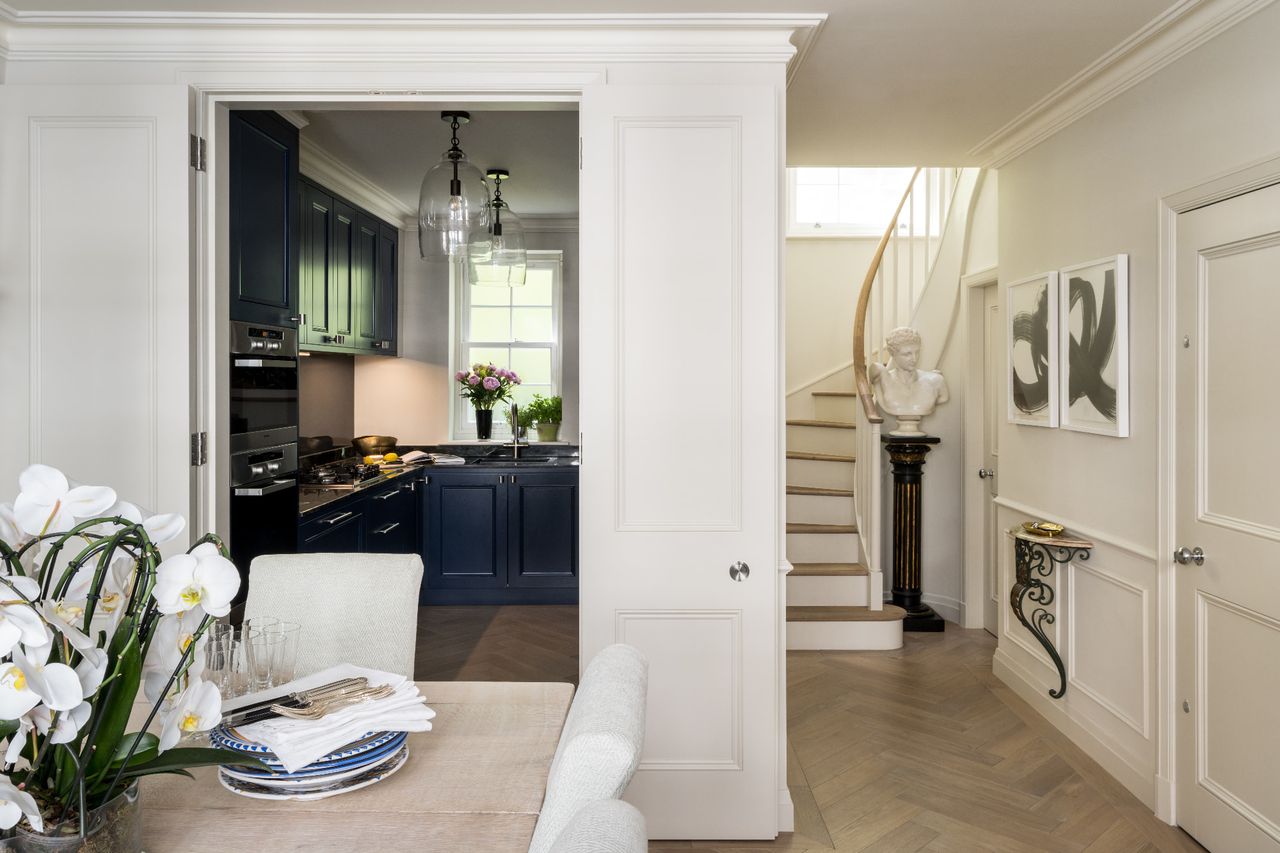 A white panelled room with light parquet flooring, a dinner table at the forefront and a blue kitchen in the background