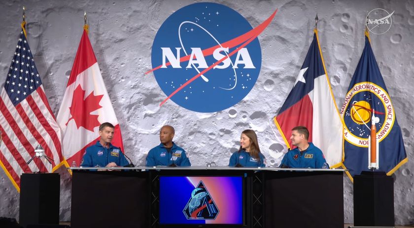 Four people wearing blue jumpsuits sit behind a large table with microphones along with four flags and a circle in the middle of the wall reading &quot;NASA&quot;