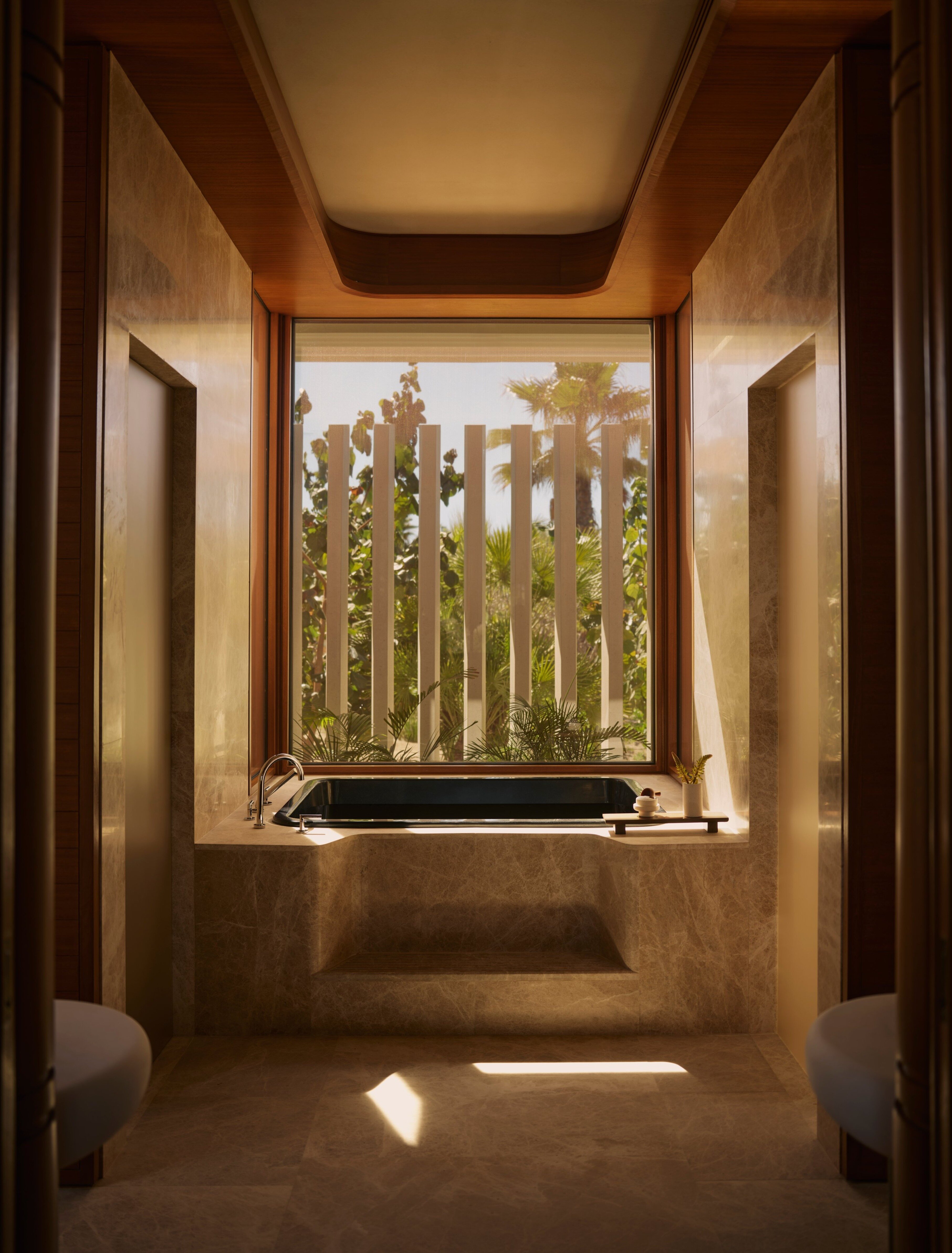 A sun-filled, stone-carved bathroom with wood detailing features a sunken bathtub that overlooks a tropical patch of greenery.