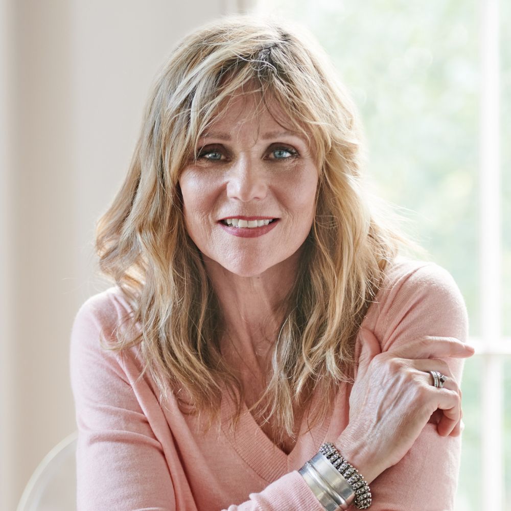Natural light frames a portrait of a woman with soft, wavy blonde hair, wearing a pale pink knit and silver bracelets.
