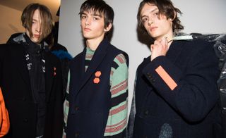 Three male models wearing looks from Off-White's collection. One model is wearing a black hooded jacket with black coat on top. Another model is wearing a blue, green and red striped top and dark blue sleeveless piece featuring two small orange hexagons. And the third model is wearing a dark blue coat with orange element and is holding a hanger with a piece of clothing