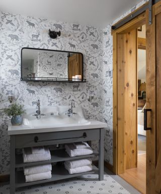 A double vanity with a white sink and two silver faucets, a dark gray base with two shelves of white folded towels, white and gray wallpaper with bear, deer, and bird illustrations on it and a black mirror