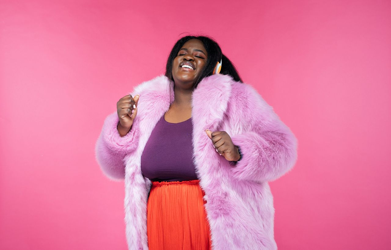 Female plus size model wearing a pink fur coat against a dark pink background.