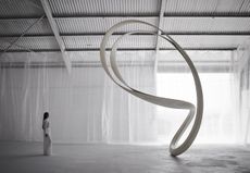A woman with long black hair in an all white longsleeve maxi dress looking ay a large white bean shaped architectural design in an all white room with high metal ceilings and white draped net like material