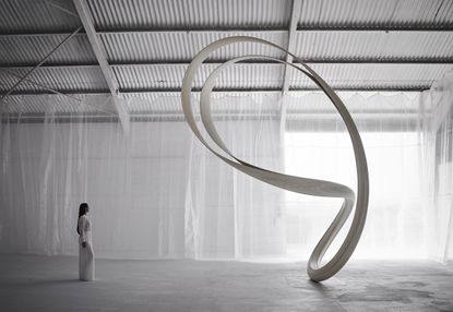 A woman with long black hair in an all white longsleeve maxi dress looking ay a large white bean shaped architectural design in an all white room with high metal ceilings and white draped net like material