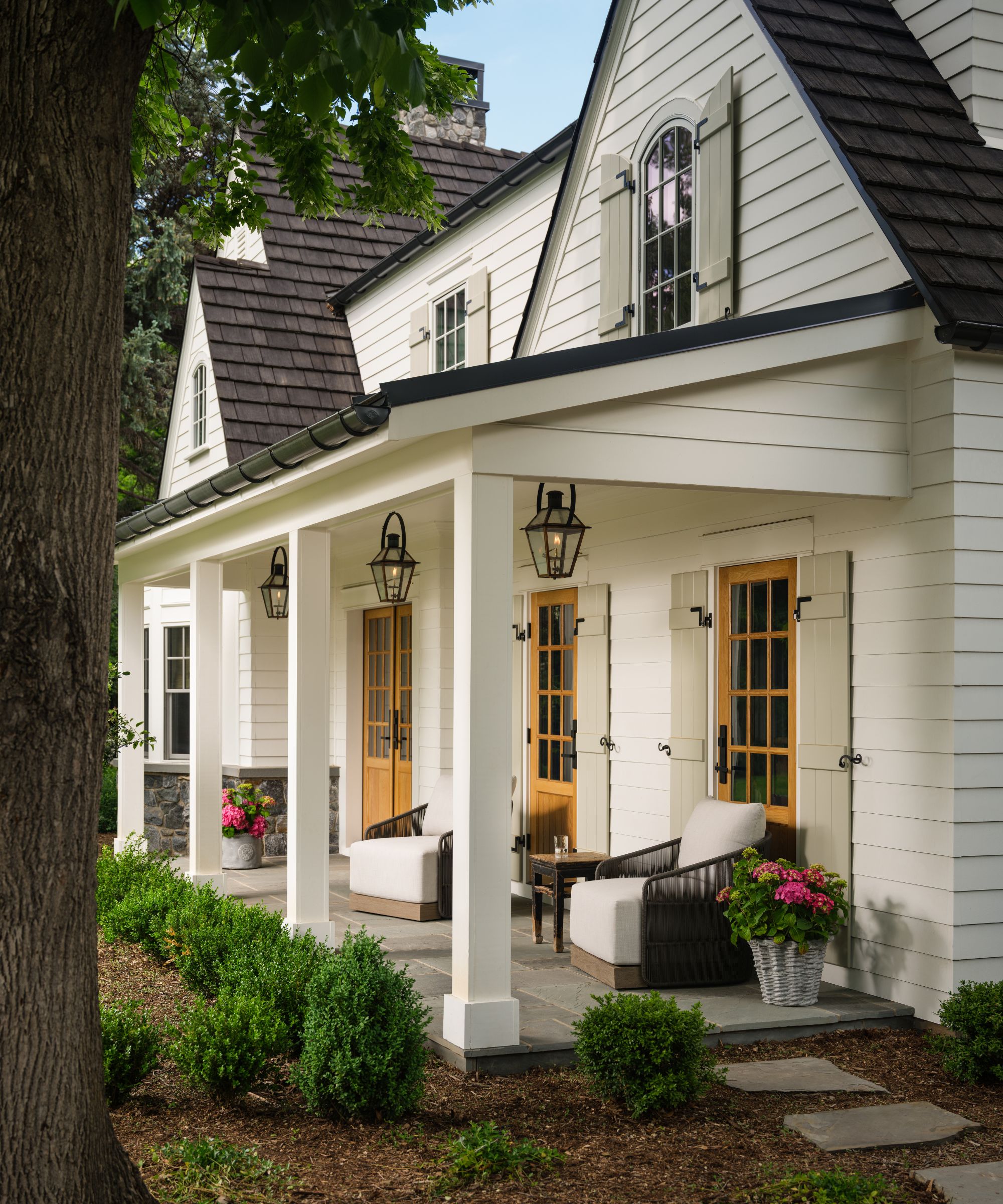 a white clad home with gabled porch, shutters and a seating area leading to a garden path and surrounded by trees
