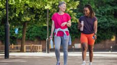Two women walk along a paved path outside, laughing and holding water bottles. Behind them we see leafy trees and lamposts.