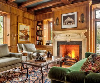 living room with fire lit taupe armchairs and green sofa and wainscot paneled walls and ceiling beams