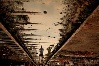 An image of artwork showing the reflection of people in standing water on the sidewalk