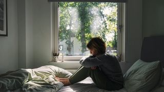 A women sitting with her arms wrapped around her legs on bed by window looking out to green trees.