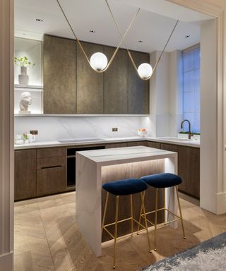 A contemporary kitchen with dark wood lower cabinetry and textured bronze upper cabinets. A white marble backsplash with subtle grey veining extends behind the countertops. The island, with a white waterfall countertop, has a wood-textured base with soft LED under-lighting