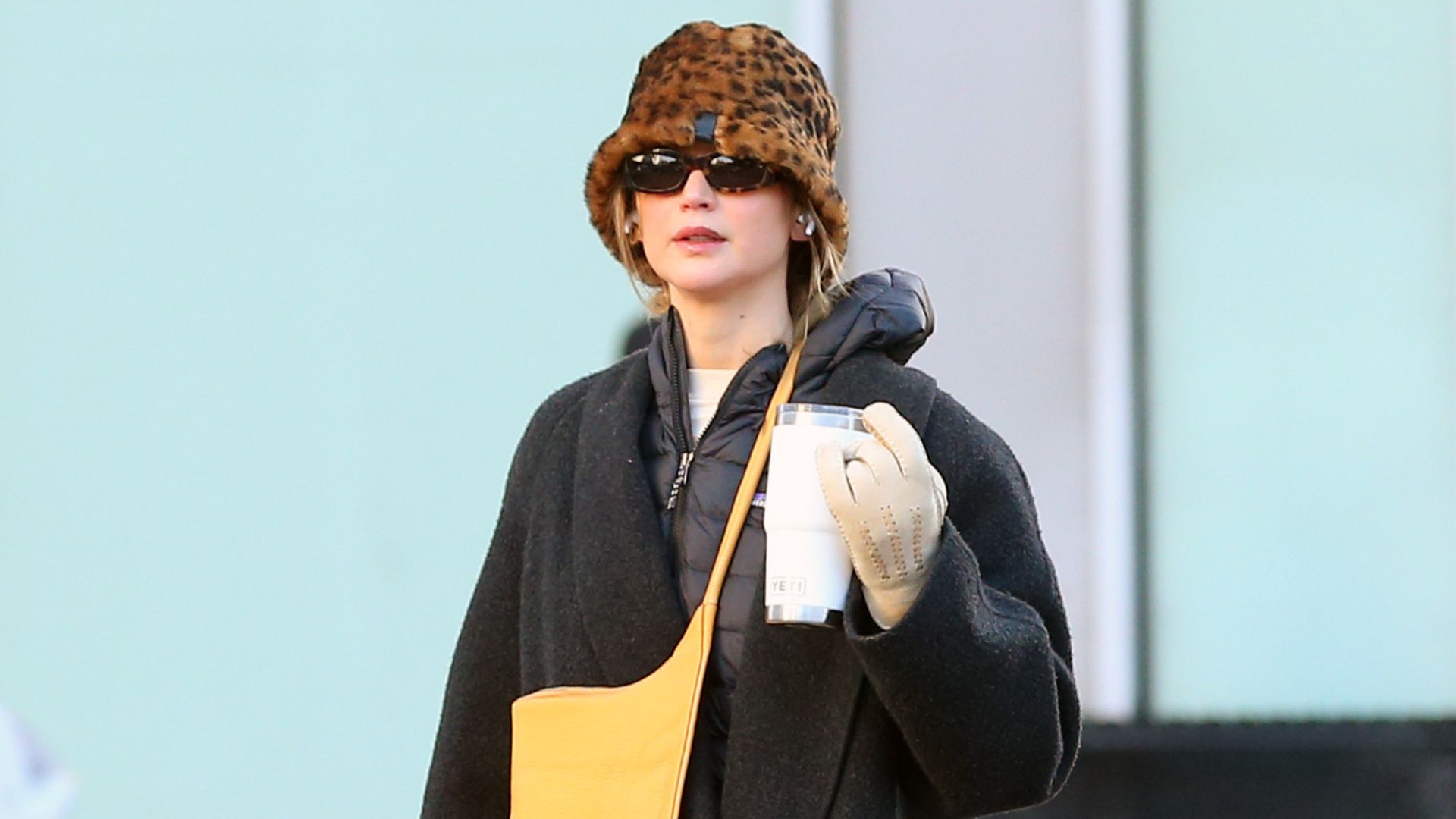 Jennifer Lawrence walks down the street wearing a black coat with Uggs, red trousers and a leopard print hat.