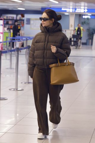 Lily James walking through Milan Malpensa Airport during Milan Fashion Week wearing a brown puffer coat and matching sweatpants, Prada sunglasses, and Prada's 2024 It bag, the buckle belt bag.