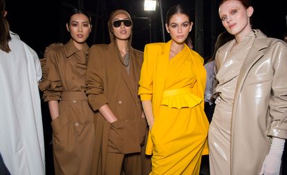 Models wear brown coats, yellow dress and blazer, sand patent leather coat and dress