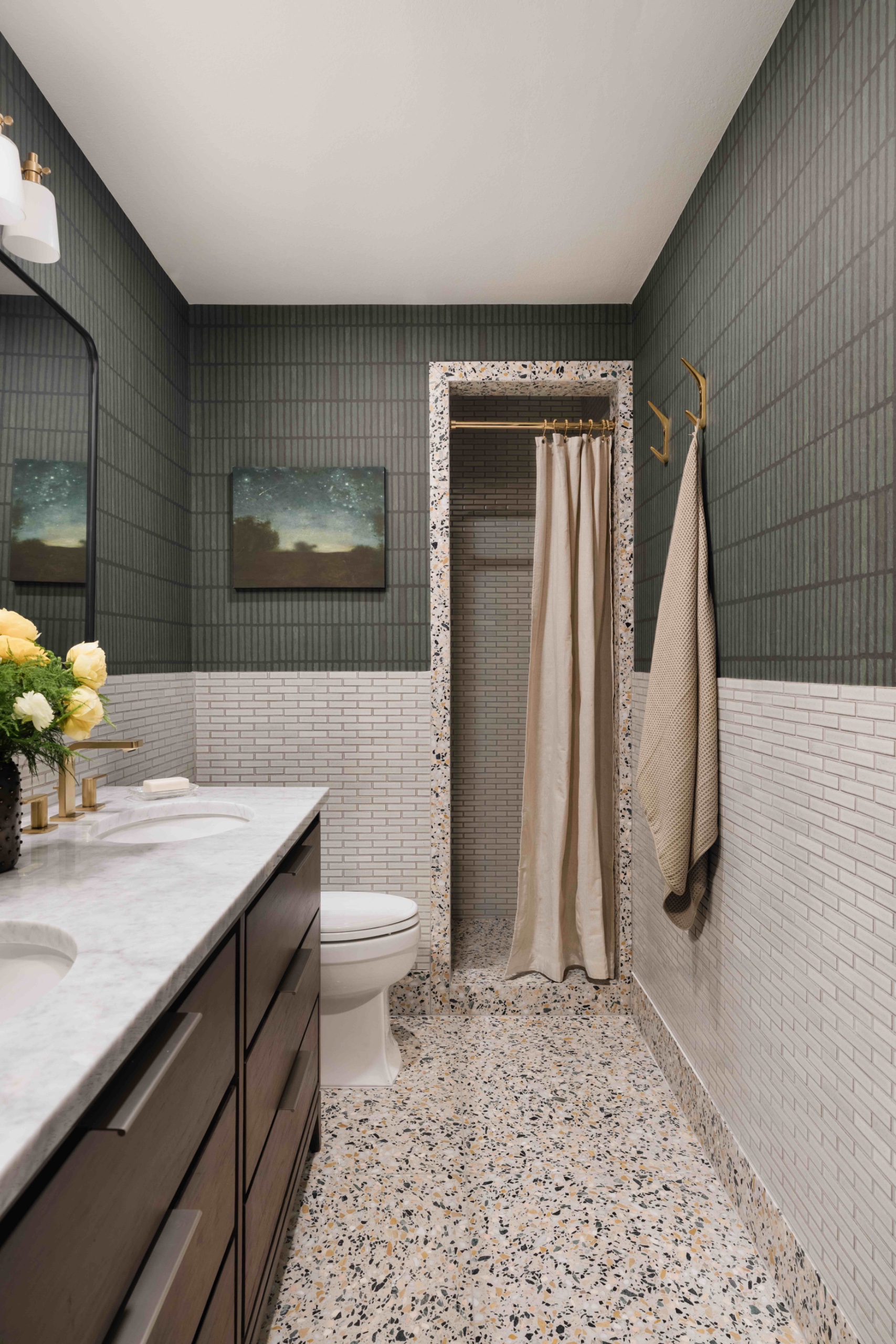 Bathroom with pale brick tiles below green grid-patterned wallpaper, a wood vanity, brass fixtures, and terrazzo flooring and shower surround.