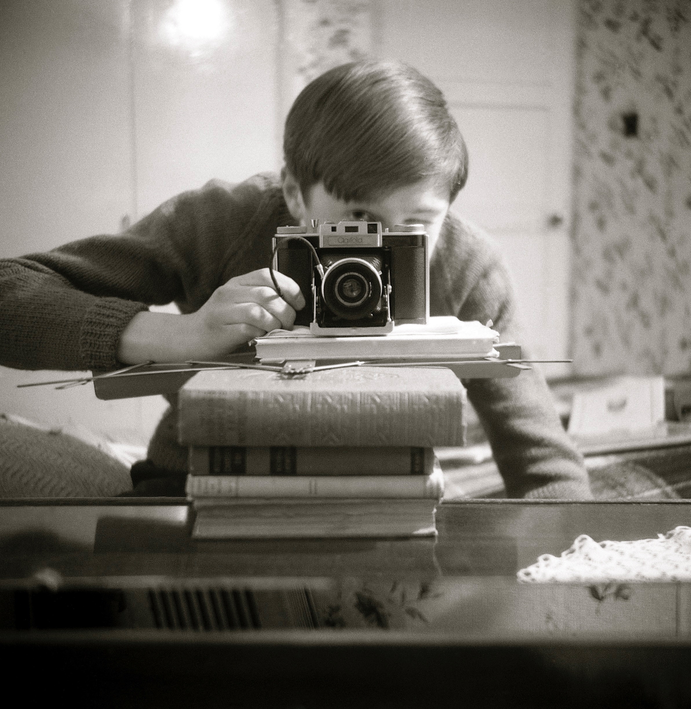Black-and-white self-portrait with an Agifold camera, on top of a stack of books obscuring the photographer's face, pointing at a mirror 