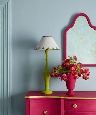 A pink mirror and dresser against a blue wall, with flowers and a lamp