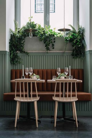 Secluded sitting area with two wooden tables and chairs, with a couch style in burnt brown that's set between walls covered in wooden boards painted in muted mint green. Plants are hanging from a large window above the sitting area.