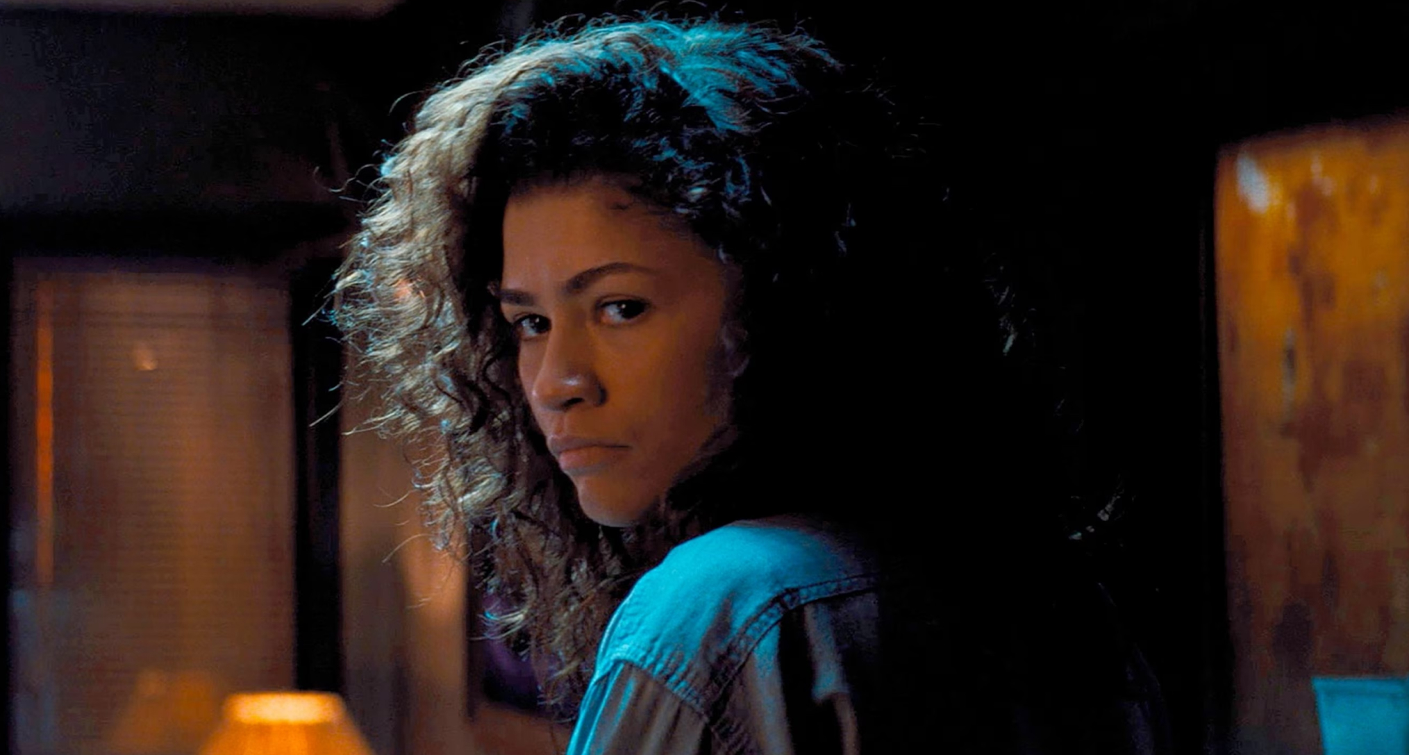 zendaya as rue looking over her shoulder in a dimly lit room in a still from euphoria season 3