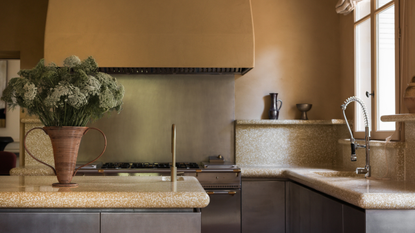A kitchen with sandy walls, brushed steel cabinets, a large vase of flowers, and a wood dining table with woven chairs
