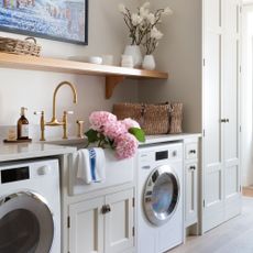 Country-style utility room with cream cabinets and a washing machine and tumble dryer either side of a white ceramic sink with pink hydrangeas in it