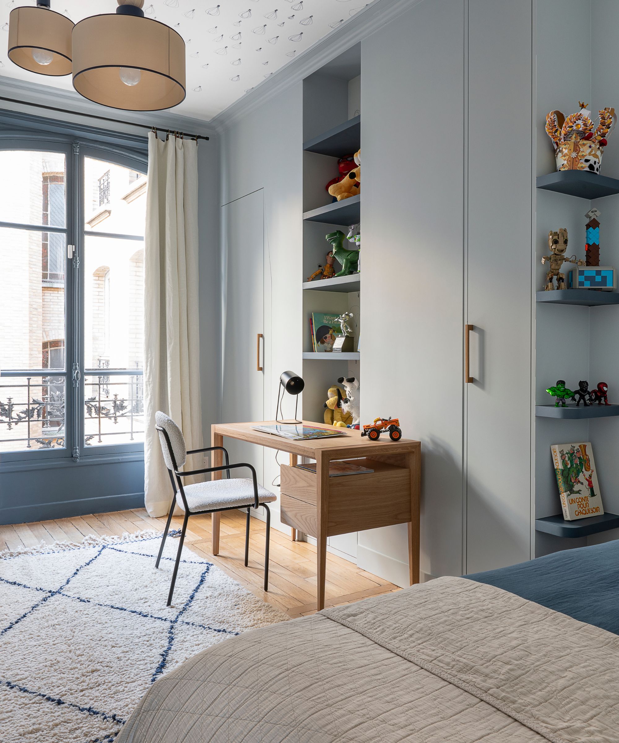 Bedroom painted in Farrow &amp;amp; Ball's Borrowed Light, a wooden desk, shelves of toys and built-in wardrobes, a glass double doors leading onto a balcony.