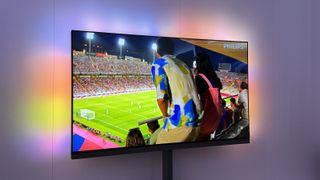 Philips OLED959 TV photographed against a white wall. On the screen is an image from a football stadium.