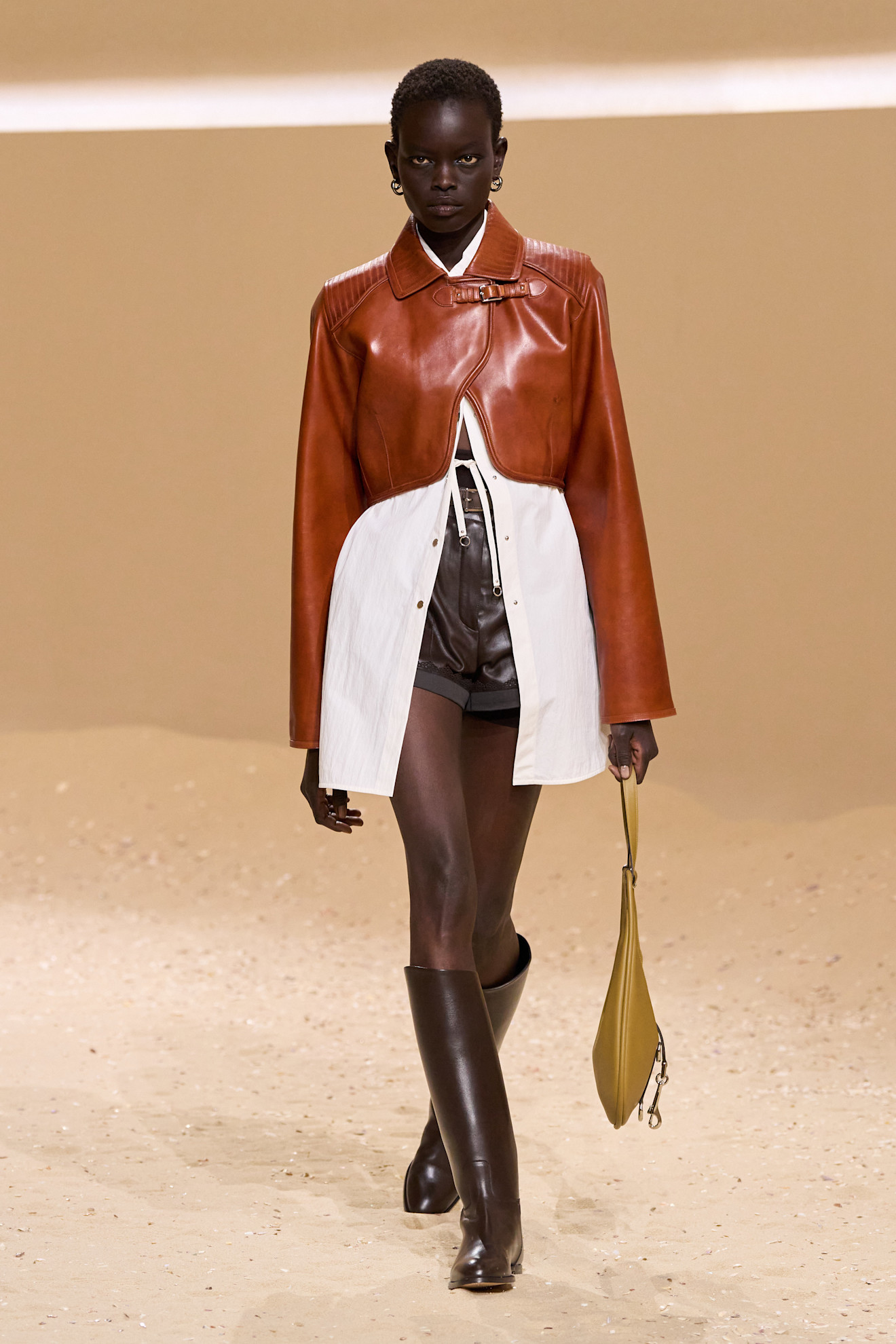 Layering staples for spring 2026 is shown in a photo from Hermes's runway show, with a model walking while wearing a tan cropped leather jacket layered on top of an oversized white button-down shirt that's unbuttoned and showing a pair of leather shorts that's styled with brown knee-high riding boots and a yellow crescent bag