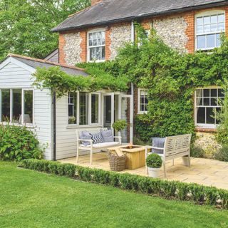 Back of a cottage covered in greenery, with patio and garden benches and part laid to lawn