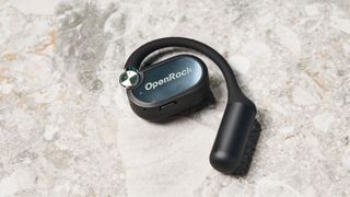 a pair of black open-ear headphones with a spring magnetic charging case photographed against a blue background