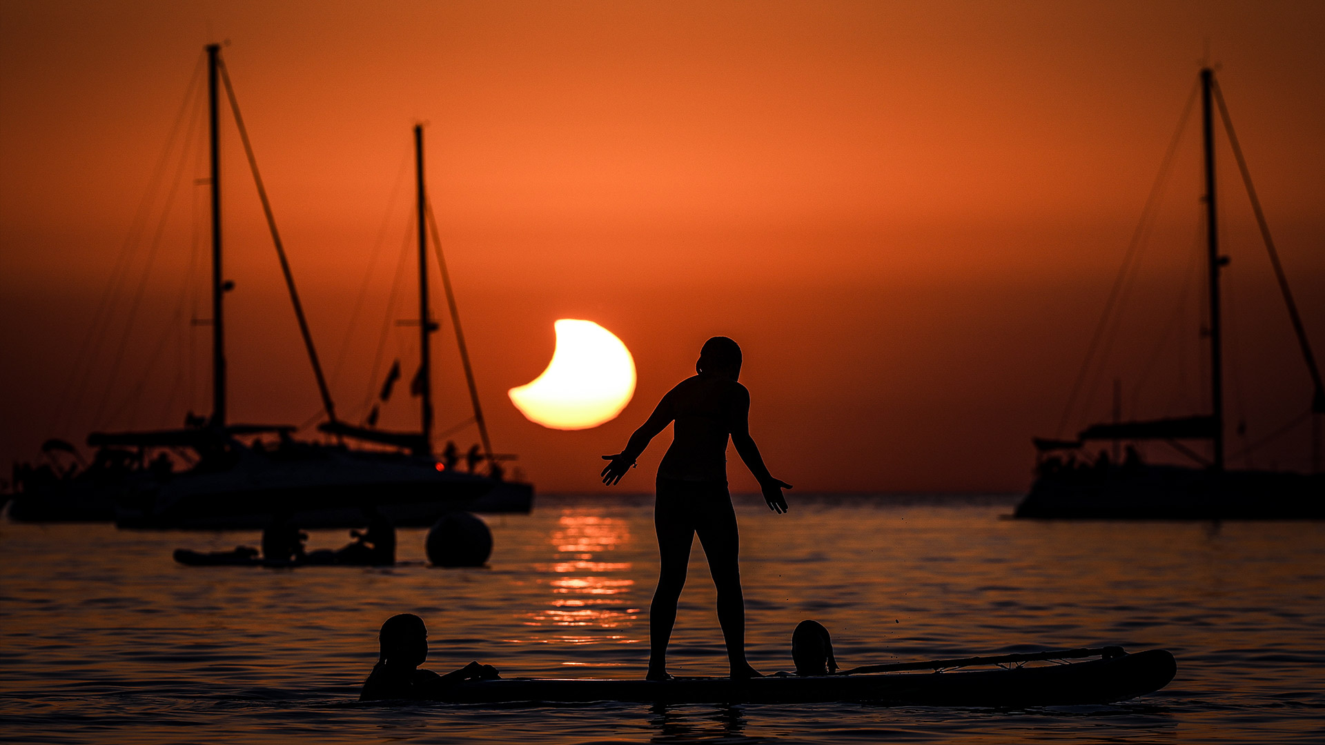 The silhouette of a woman standing on a paddle board next to two sail boats with the solar eclipse in front of her.
