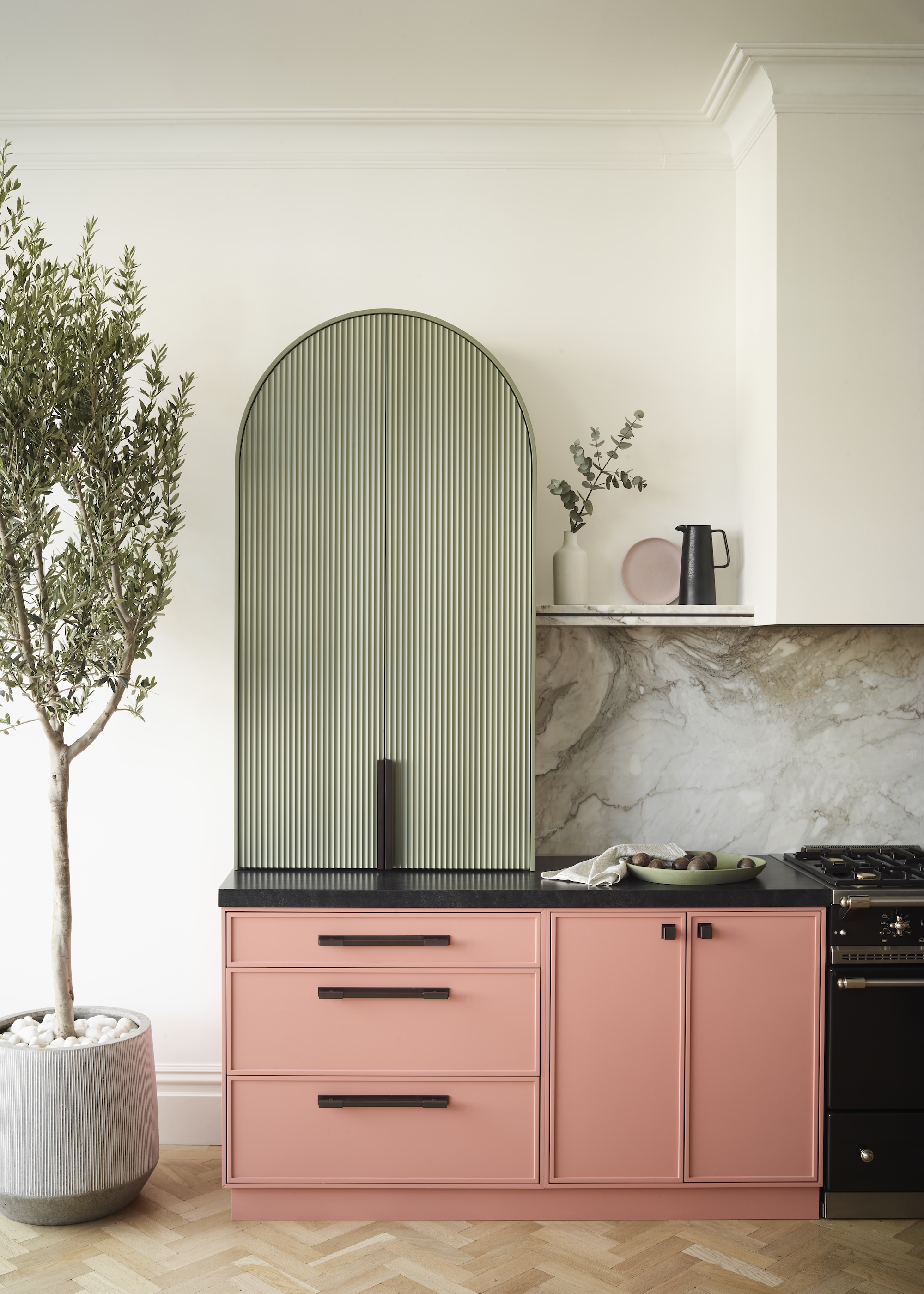 A kitchen showing salmon pink base cabinets with a sage green curved wall cabinet with fluted detail and black worktops