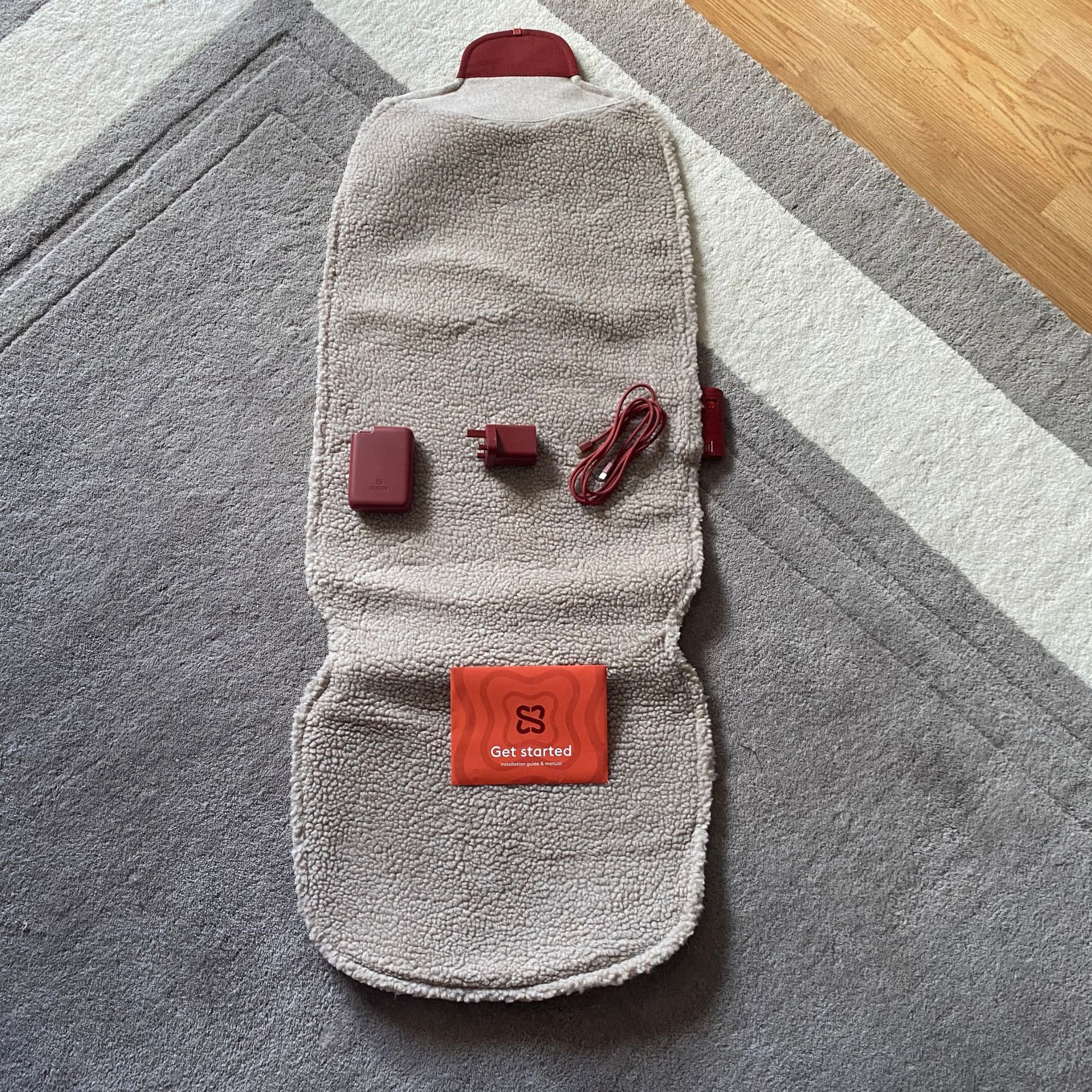 An unrolled Stoov Big Hug3 on a neutral rug, with the battery, plug and cable laid on top