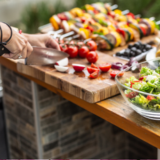 Garden table full of food ready for a barbeque party with a salad in the front and a woman cutting vegetable aside.