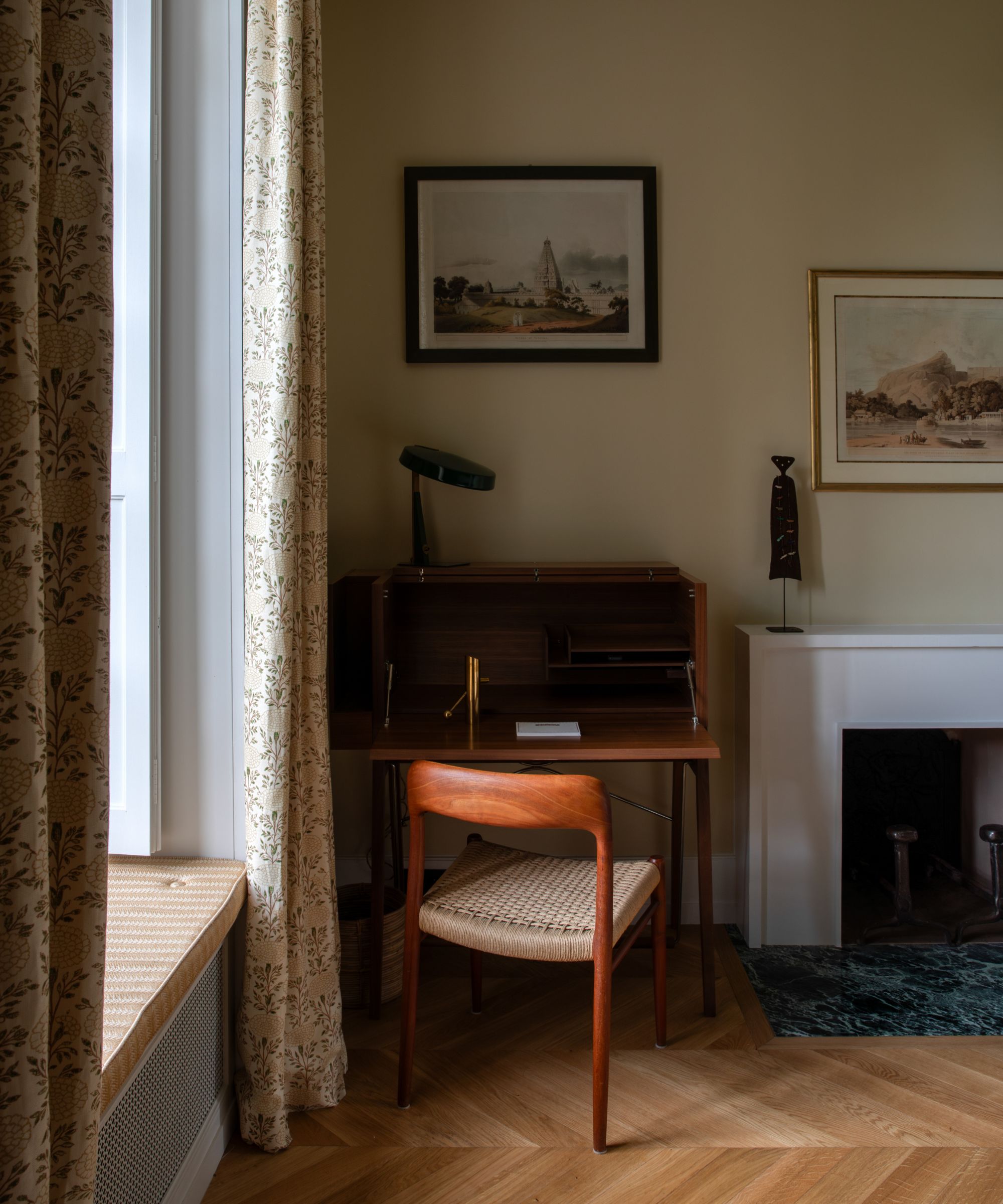 A small wooden writing desk with a matching chair in the corner of a neutral painted room with wooden herringbone flooring.