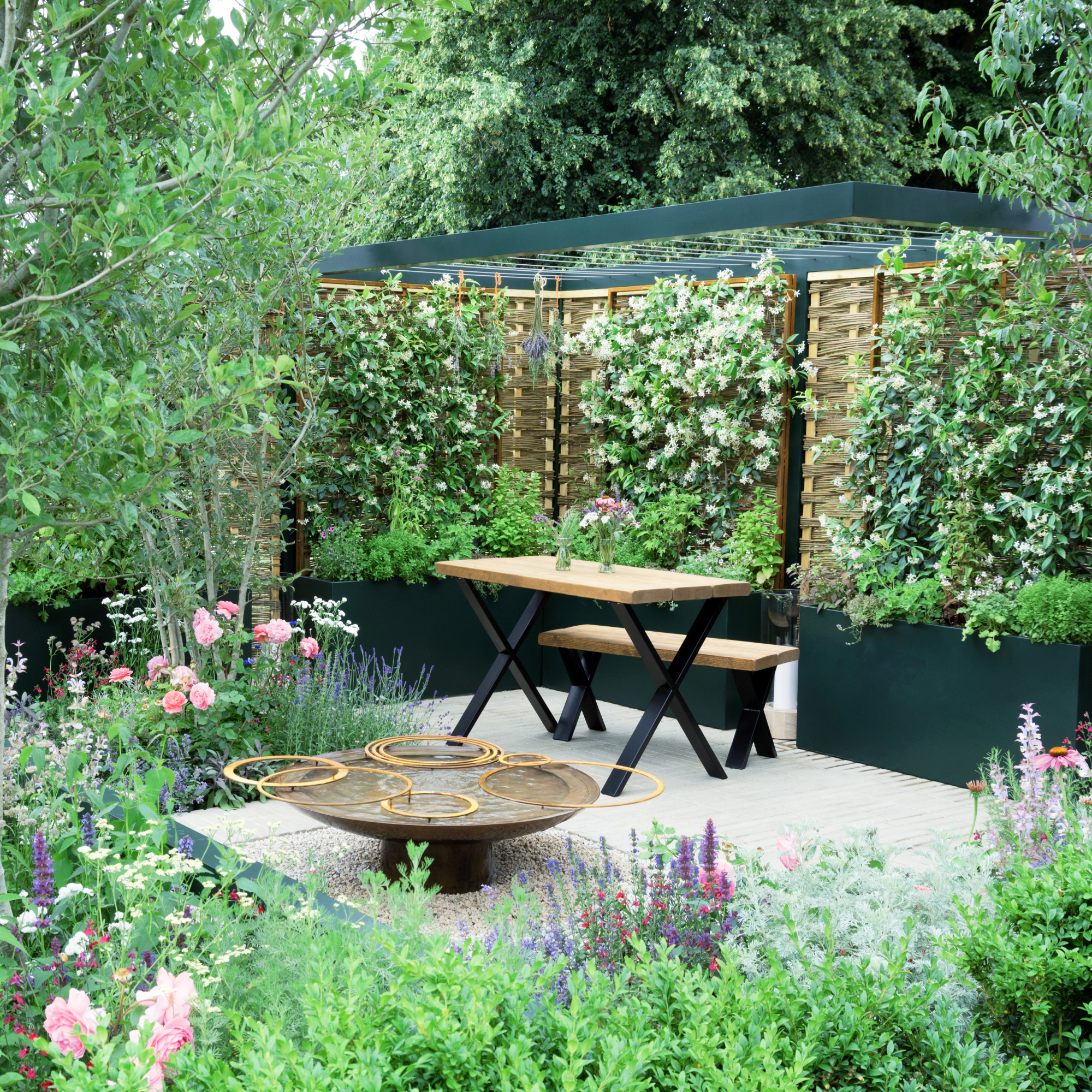 small garden with plenty of vibrant perennials planted in containers, bench and outdoor table, and water feature