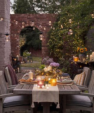 Ambient outdoors dining set-up with festoon lighting overhead, twinkling fairy lights in bushes, and glowing candles staggered along pretty tablescape with lilac florals and striped runner.